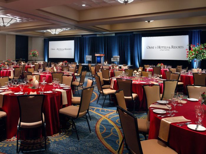 The Grand Ballroom at the Omni Charlotte Hotel. The event decor is dressed with red tablecloths at multiple tables in the grand ballroom. The LED Screens on the back wall have the Omni's branding on it.