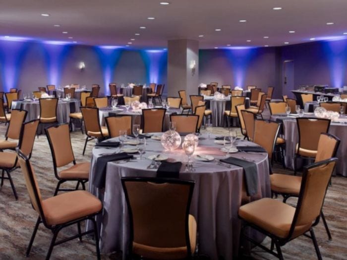 Th Cypress Room at The Omni Charlotte Hotel. The Event has blue lighting on the walls and multiple round tables throughout it. The tables are set with grey tableclothes and orange chairs.